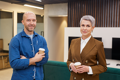 Waist up portrait of two mature people holding coffee cups standing in modern office interior and smiling at camera, copy space
