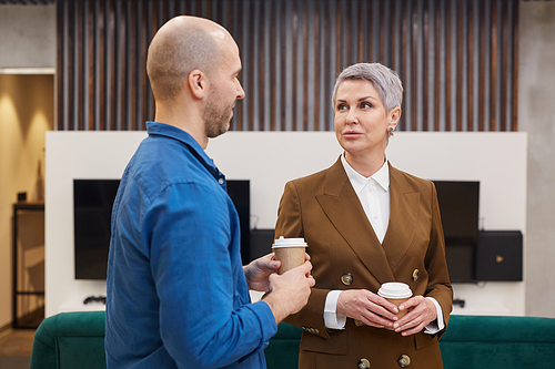 Waist up portrait of modern mature businesswoman talking to colleague and holding coffee cup during break in office lobby