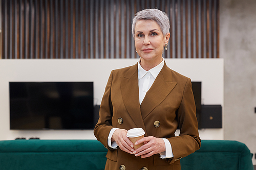 Waist up portrait of modern mature businesswoman holding coffee cup and  while standing in office lobby, copy space