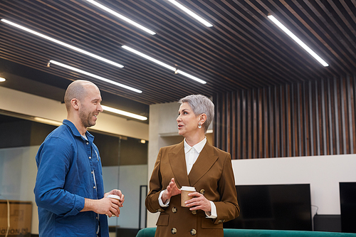 Wide angle waist up portrait of modern mature businesswoman talking to colleague and holding coffee cup during break in office lobby, copy space