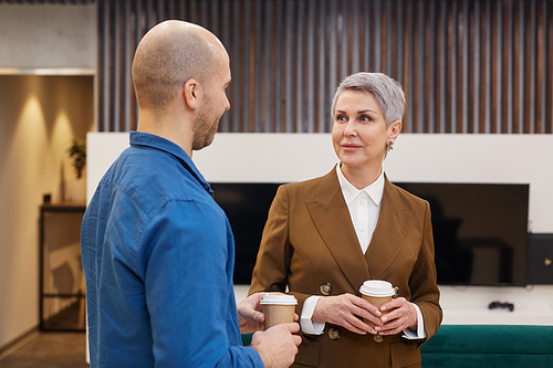 Waist up portrait of modern mature businesswoman chatting with colleague and holding coffee cup during break in office lobby