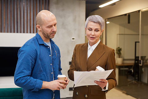 Waist up portrait of modern mature businesswoman holding documents and talking to colleague while standing in office lobby
