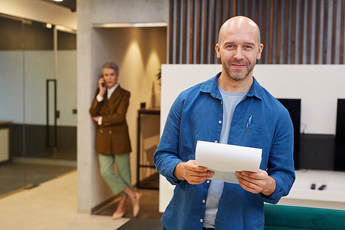 Waist up portrait of bald mature man smiling cheerfully at camera while holding contract in office lobby, satisfied client concept, copy space