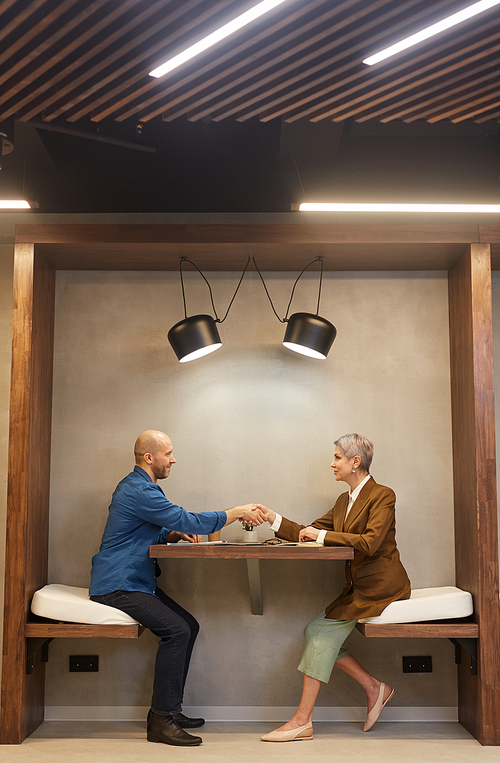 Side view full length portrait of two people shaking hands while sitting at table in indoor cafe during business meeting, copy space
