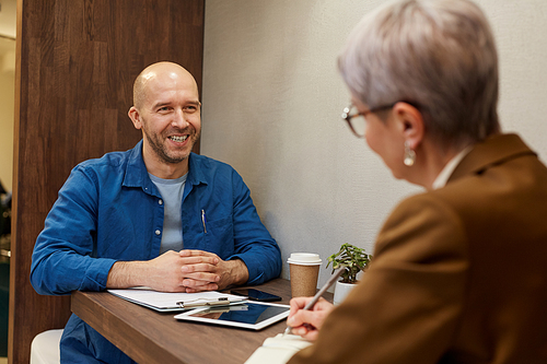 Portrait of mature bald man smiling happily while talking to business manager during meeting at cafe table, copy space