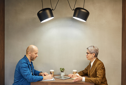 Minimal side view portrait of mature bald man writing on clipboard while discussing contract with business manager during meeting at cafe table, copy space