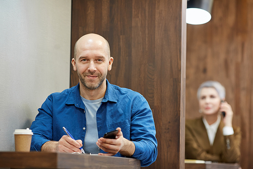 Portrait of bald adult man smiling happily at camera while working at cafe table and holding smartphone, copy space