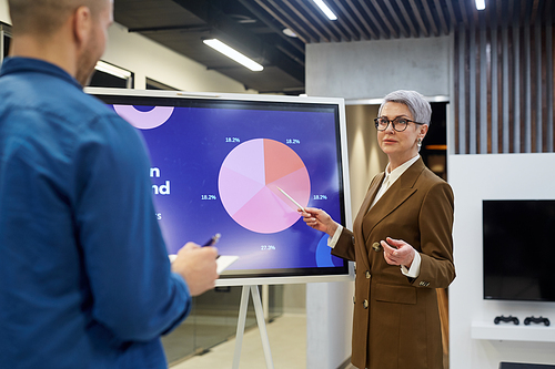 Portrait of modern mature businesswoman pointing at pie chart while giving presentation for employees or clients in office hall, copy space