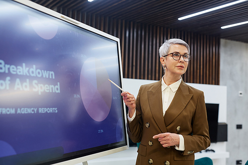 Waist up portrait of successful mature businesswoman pointing at pie chart while giving presentation on marketing, copy space