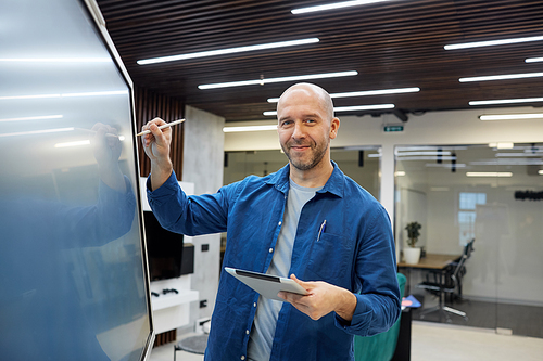Waist up portrait of bald adult man smiling at camera while using digital board during presentation or training seminar in office, copy space