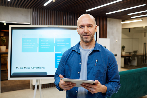 Waist up portrait of bald adult man smiling at camera and holding tablet while standing against digital board during marketing presentation, copy space