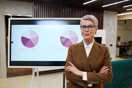 Waist up portrait of modern mature business manager  while standing with arms crossed against digital board during marketing presentation, copy space
