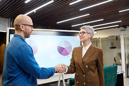 Waist up portrait of mature female manager shaking hands with client or employee while standing against presentation board, copy space