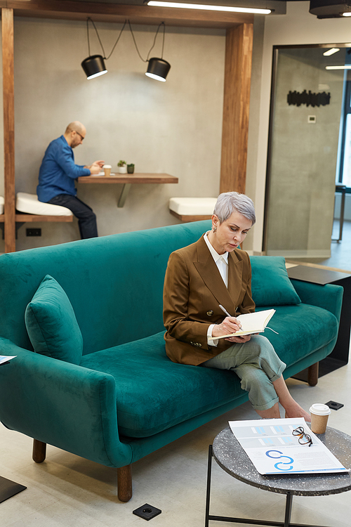 Full length portrait of modern mature businesswoman writing in note book while sitting on couch in designer office interior, copy space