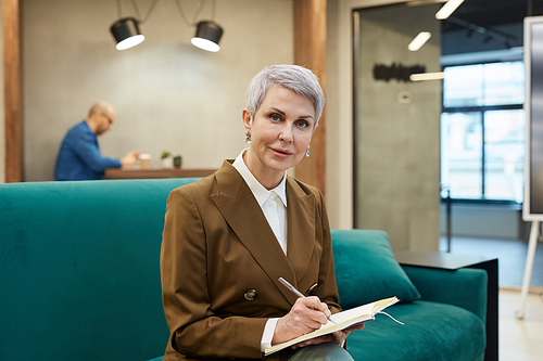 Portrait of elegant mature businesswoman writing in note book and  while sitting on couch in modern office interior, copy space