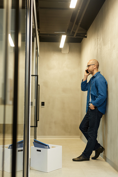 Full length side view at bald adult man speaking by smartphone while leaning on wall in minimal office hall, copy space