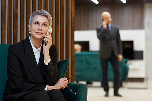 Portrait of successful mature woman speaking by phone in modern office interior, copy space