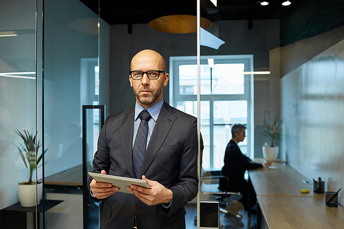 Waist up portrait of successful bald businessman standing in modern office interior holding tablet, copy space