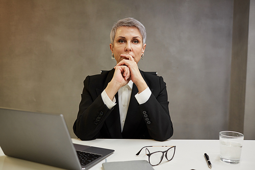 Minimal portrait of elegant mature businesswoman looking away pensively while sitting against grey wall at workplace, copy space