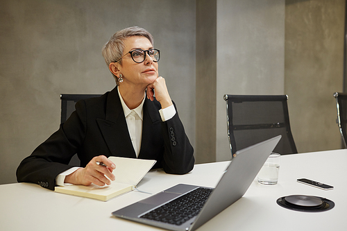 Minimal portrait of successful mature businesswoman looking away pensively while sitting against grey wall at workplace, copy space
