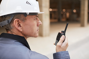 Side view portrait of mature worker speaking by walkie-talkie while supervising work at construction site or in industrial workshop, copy space