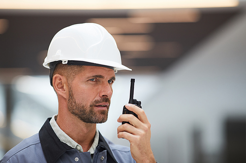 Close up portrait of mature worker speaking by walkie-talkie while supervising work at construction site or in industrial workshop, copy space