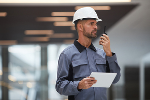 Waist up portrait of handsome mature worker speaking by walkie-talkie while supervising work at construction site or in industrial workshop, copy space