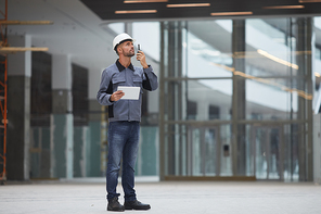 Full length portrait of mature worker speaking by walkie-talkie while supervising work at construction site or in industrial workshop, copy space