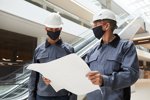 Waist up portrait of two construction workers wearing masks and discussing plans while standing in shopping mall or office building