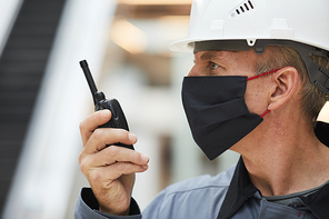 Side view portrait of mature worker wearing mask and speaking by walkie-talkie while working on industrial site, copy space