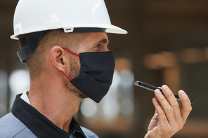 Side view portrait of mature worker wearing mask and recording voice message while working on industrial site, copy space