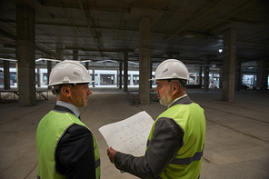 Back view at two business people wearing hardhats and holding plans while standing at construction site, copy space