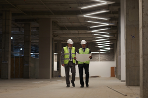 Full length portrait of two business people wearing hardhats and holding plans while walking towards camera at construction site, copy space