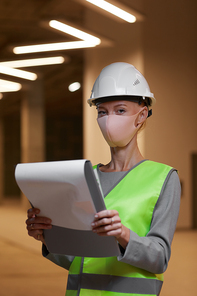 Vertical waist up portrait of mature female worker wearing mask and  while standing with clipboard at construction site indoors