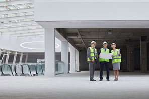 Wide angle portrait of three successful business people wearing hardhats and looking up while standing at construction site indoors, copy space