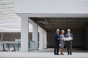 Wide angle portrait of three successful business people wearing hardhats and holding plans while inspecting construction site indoors, copy space