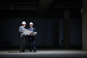 Dramatic full length portrait of two mature business people wearing hardhats and holding plans while standing in dark at construction site lit by harsh lighting, copy space