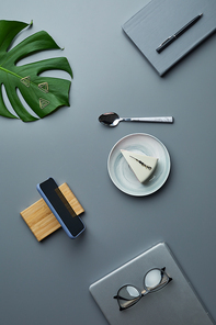 Minimal above view flat lay of cheesecake and business accessories over grey workplace background with tropical leaf, copy space