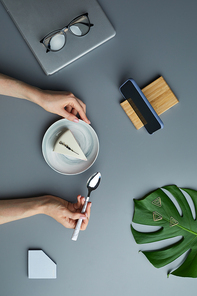 Minimal above view flat lay of woman eating cheesecake over grey workplace background with tropical leaf and business accessories, copy space