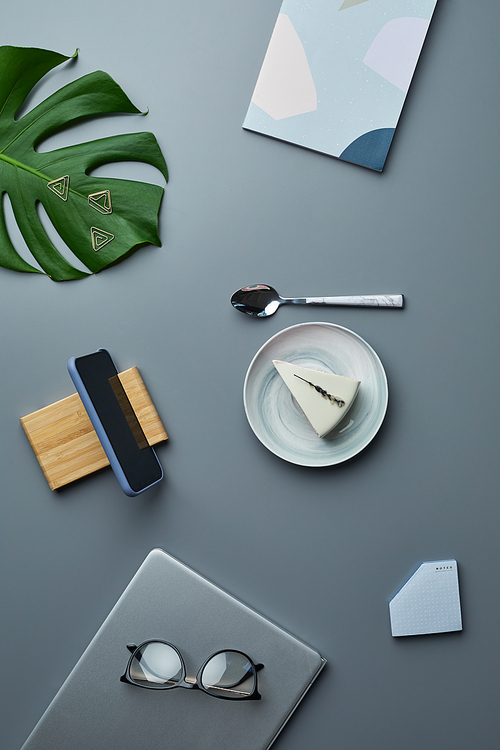 Minimal top view flat lay of cheesecake and business accessories over grey workplace background with tropical leaf, copy space