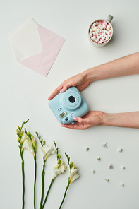 Minimal background composition of female hands holding instant camera over while table background with floral accents and cup of cocoa, copy space