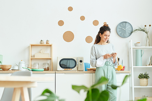 Happy young woman wearing stylish outfit standing in her kitchen texting message to someone using her smartphone