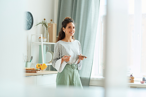 Stylish young woman wearing casual outfit standing in kitchen taking part in online meeting using her smartphone and earphones