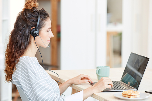 Modern young Caucasian woman wearing headset working on laptop at home, horizontal side view shot