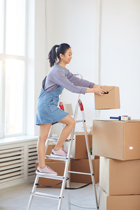 Vertical full length portrait of young Asian woman stacking cardboard boxes while standing on ladder in new house, moving in concept, copy space