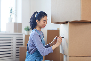Side view portrait of smiling Asian woman writing on cardboard boxes labeling them for moving out to new house, copy space