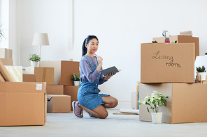 Full length portrait of young Asian woman organizing moving in process while sitting on floor next to cardboard boxes and holding planner, copy space