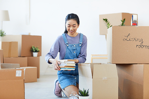 Full length portrait of young Asian woman packing books to cardboard boxes and smiling happily excited for moving to new house or dorm, copy space