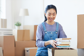 Portrait of young Asian woman packing books to cardboard boxes and smiling happily excited for moving to new house or dorm, copy space