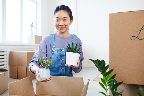 Portrait of young Asian woman packing plants to cardboard boxes and smiling at camera while moving to new house or apartment, copy space